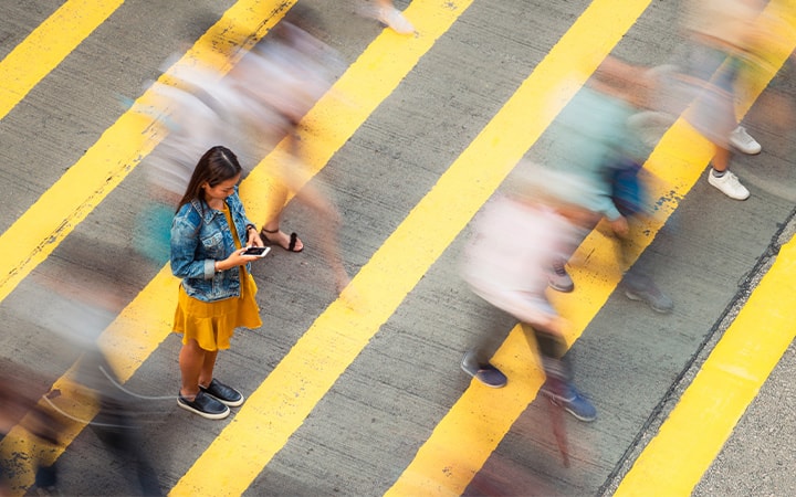 A woman, stationary on a crosswalk as people move past her, looking down to her smartphone