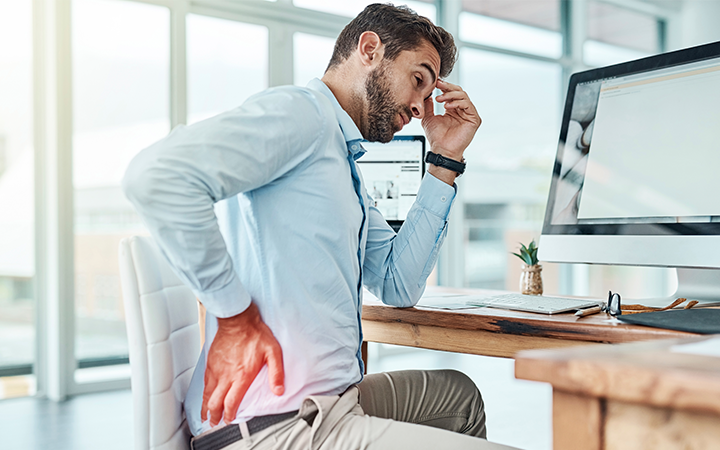 man sitting at desk with hand on his back as if in pain