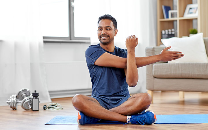 man seated with crossed legs on exercise mat stretching arm across his body