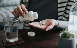 Close up of young Asian woman holding a pill bottle, pouring pills into palm of hand, with a glass of water on the side.