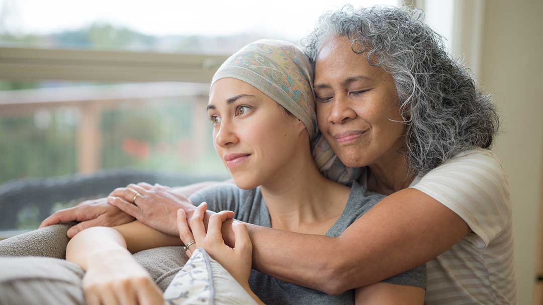 woman in 50s embracing her mid-20s daughter on couch who is fighting cancer