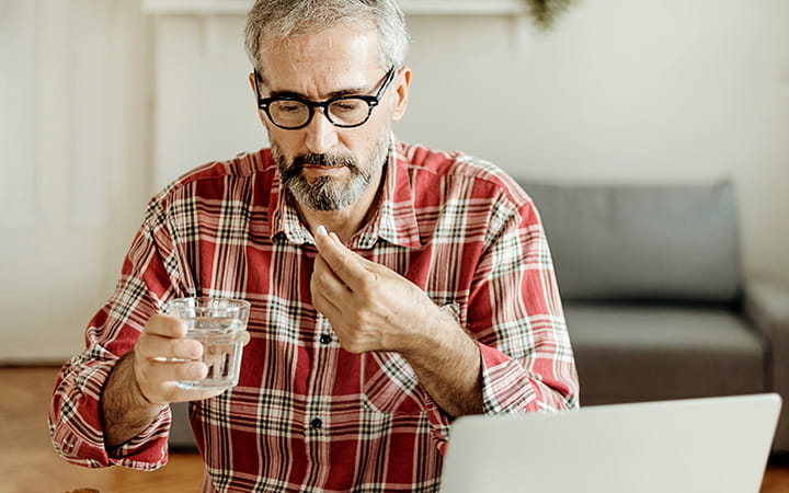 middle aged man taking aspirin with a glass of water in front of laptop
