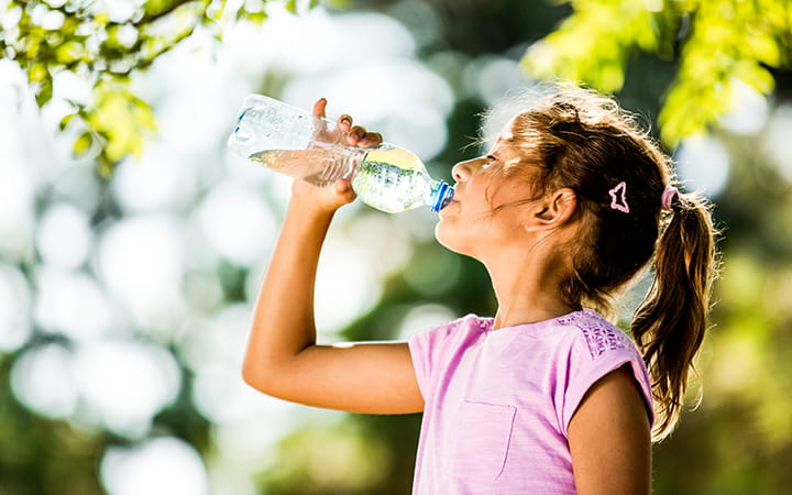 young girl in pink top drinking glass of water with trees in the background