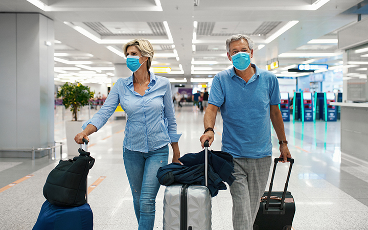 masked couple in blue walking through airport with roller luggage