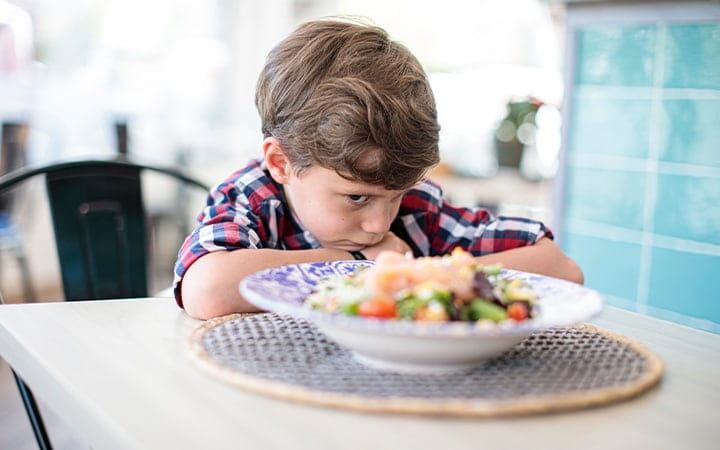 An angry child seated at the table glares at his food