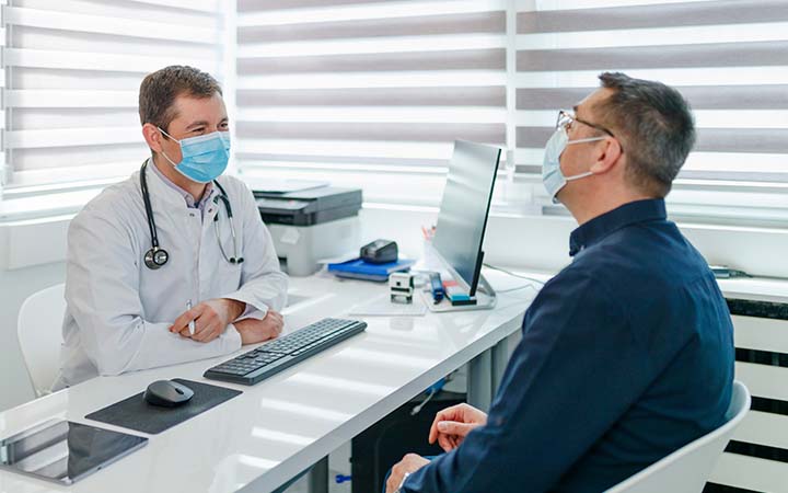 male masked doctor talking with masked male patient over a white desk