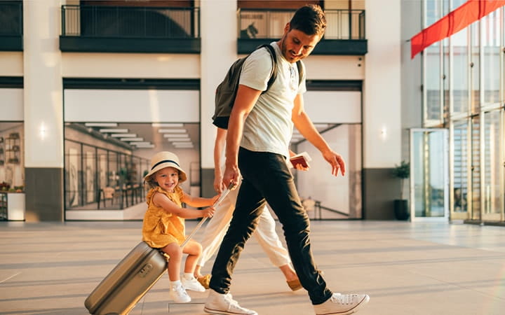 Cheerful husband and his anonymous wife walking with their little girl sitting on luggage at the airport