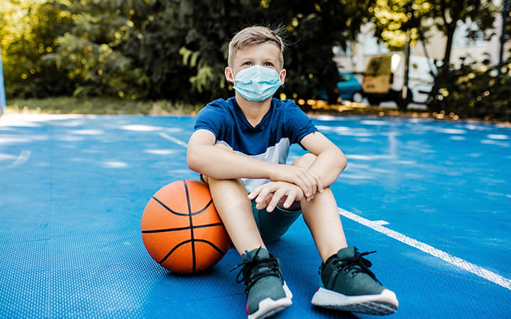 young boy with mask sits on basketball court with a basketball