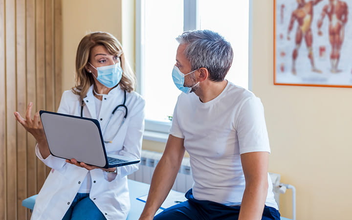 female doctor with laptop in mask talking with older male patient in mask