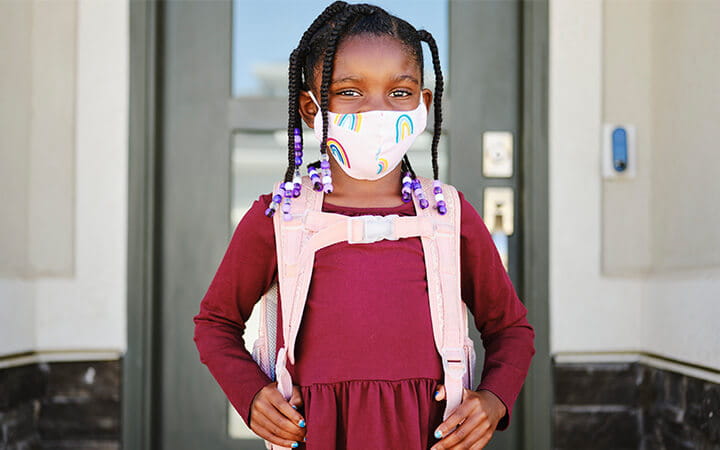 An elementary aged school student leaving her home to go back to school, wearing a mask for protection against infectious disease