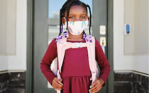 An elementary aged school student leaving her home to go back to school, wearing a mask for protection against infectious disease