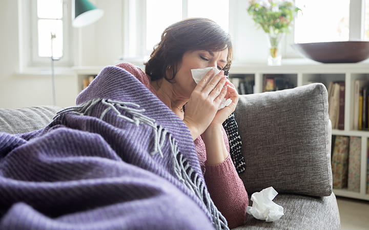 woman wrapped in blanket on a couch blowing her nose
