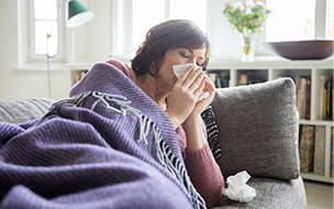 woman wrapped in blanket on a couch blowing her nose