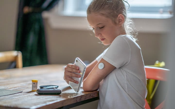 A young girl reads her blood sugar level by placing her cellphone next to her pump in her arm as she sits at a dining table
