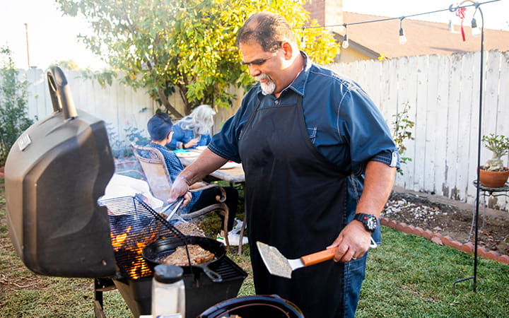 older man wearing blue apron tends to grill with both hands holding implements