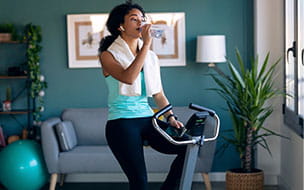 woman on stationary cycle drinking water from a bottle with towel around her neck