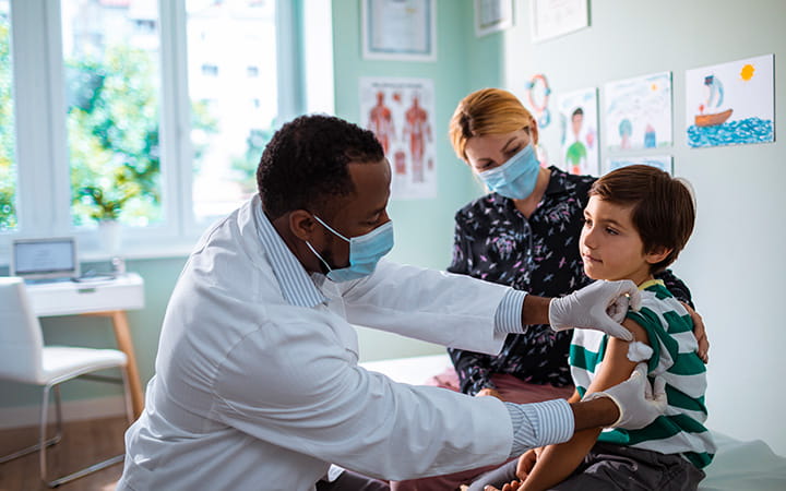 masked male doctor giving vaccine to young boy as masked mother looks on
