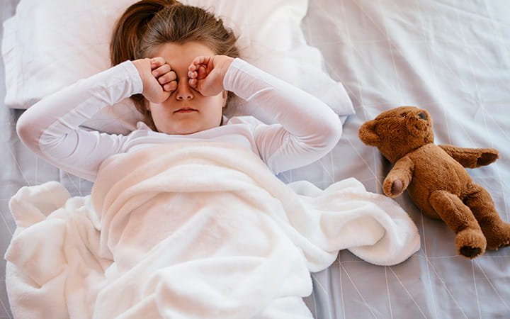 child lying in bed with teddy bear rubbing eyes