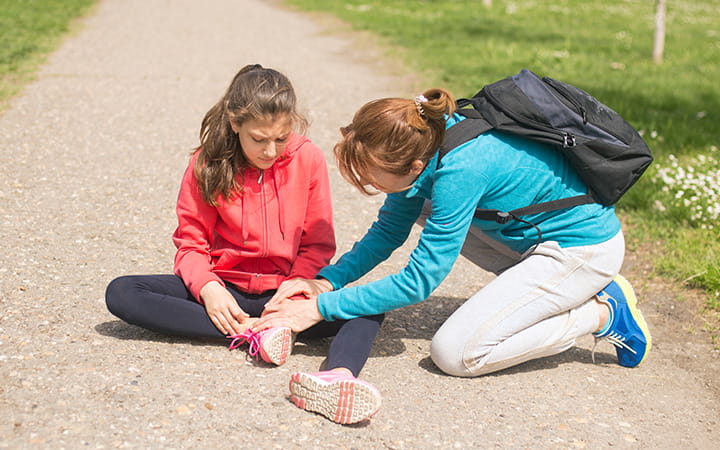 girl with ankle injury sits on path with other girl helping
