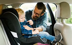 father buckling young child into car seat