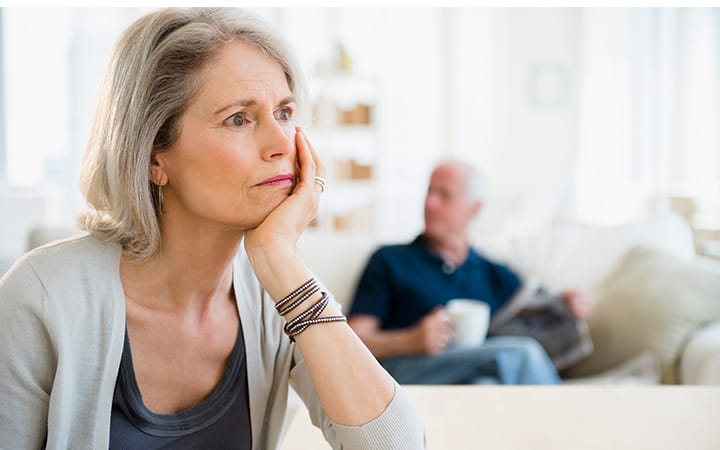 older woman resting chin in hand with man on couch in background