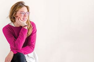 girl in hot pink sweater and glasses sits cross-legged with head in hands