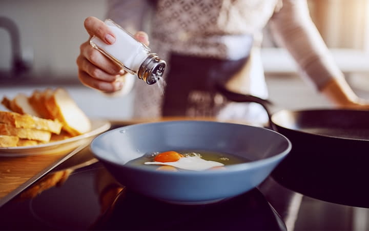 Woman sprinkling salt on an egg cooking in a frying pan.