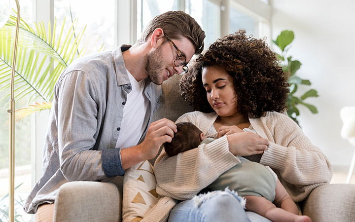 A young woman breastfeeds her infant son as her husband smiles at the baby