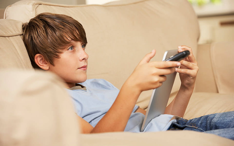 Young boy sitting on sofa at home using tablet computer whilst watching television
