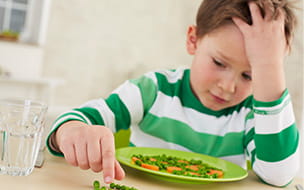 Young boy playing with his peas at the table because he doesn't want to eat them.