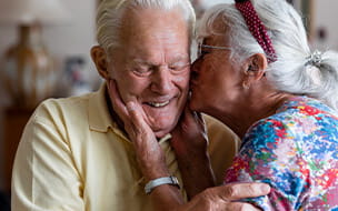 Senior couple, at home, wife kissing husband's cheek while he laughs with his eyes closed