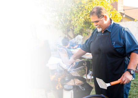 A older man wearing blue apron tends to grill with both hands holding implements