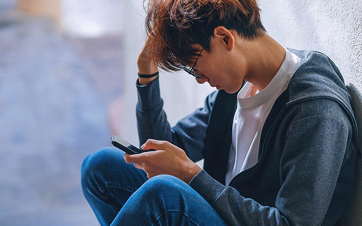 An upset teenage boy, dressed in casual outfit, sits on a stairway reading on his smartphone with a backpack beside him, while dealing with the issue of cyberbullying