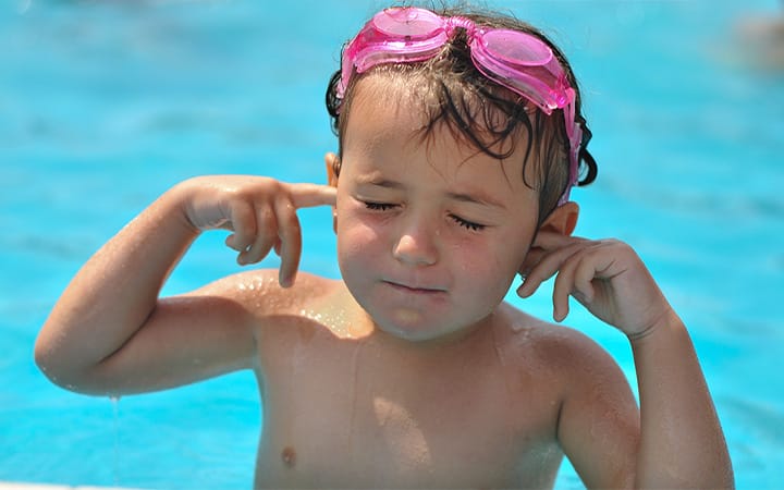 A little girl in a pool wearing swimming goggles holding her fingers in her ears