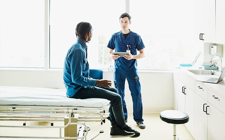A male nurse holding a digital tablet while in discussion with a patient in an exam room 