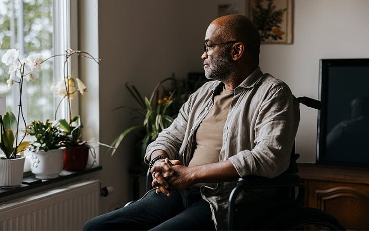 A thoughtful senior man sitting with hands clasped on wheelchair near window at a nursing home