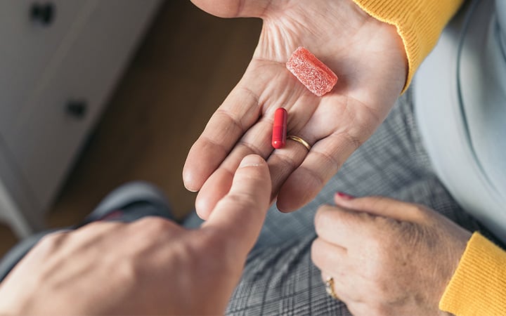 A middle-aged woman in a yellow sweater sitting on her bed holding a red pill and a jelly bean