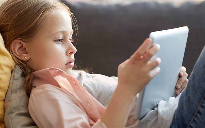A young girl uses a digital tablet while lying on sofa, watching cartoons or browsing the internet