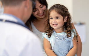 Little girl of Hispanic decent, sits on her mothers lap, on an exam table both smiling as they talk with the male doctor.