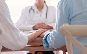 A close up of a couple holding hands resting on a desk across from a doctor