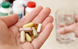 Close up of a left hand holding a variety of vitamins, supplements and herbal remedies, while their right hand holds a glass of water.