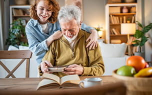 Granddaughter hugging her grandfather while he reads a book in the living room at home