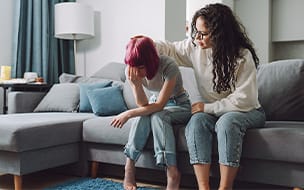 Teenage girl covering her face and crying on a couch, while her mother strokes her head