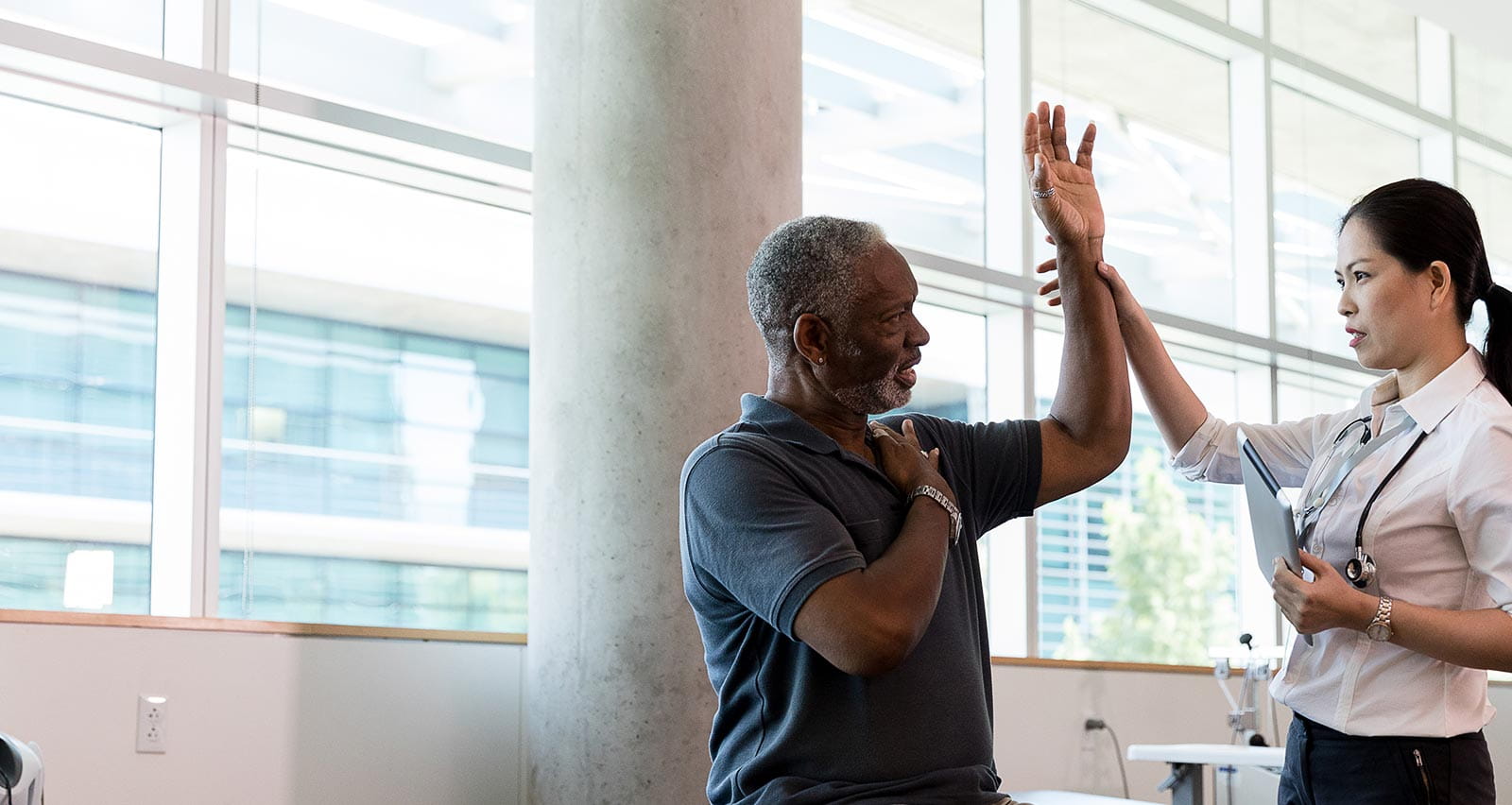 Confident female orthopedic doctor has a senior male patient lift his hand over his shoulder, testing his range of motion in his arm