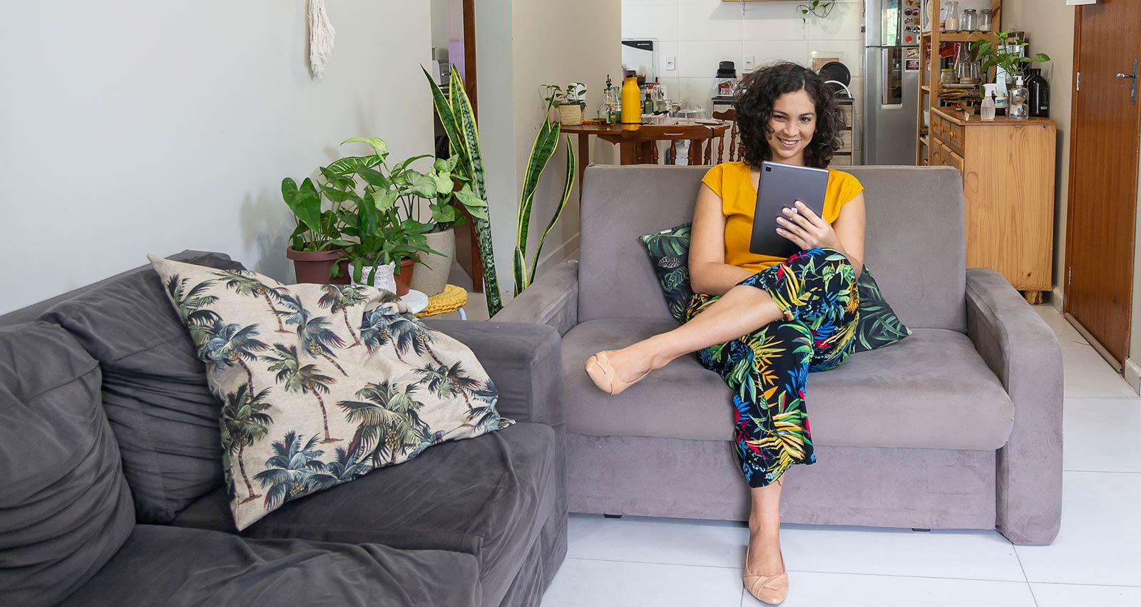 Latin woman over thirty with curly hair and yellow blouse and using tablet for a video call on a sofa in a living room with plants