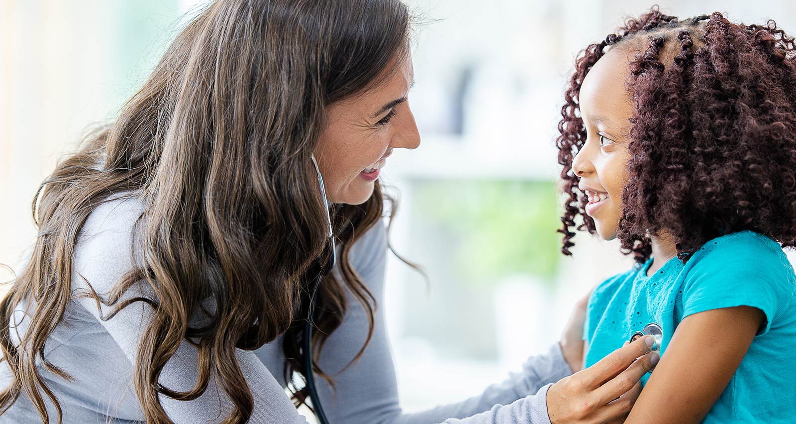 A little girl sits up on an exam table as her doctor listens to her heart