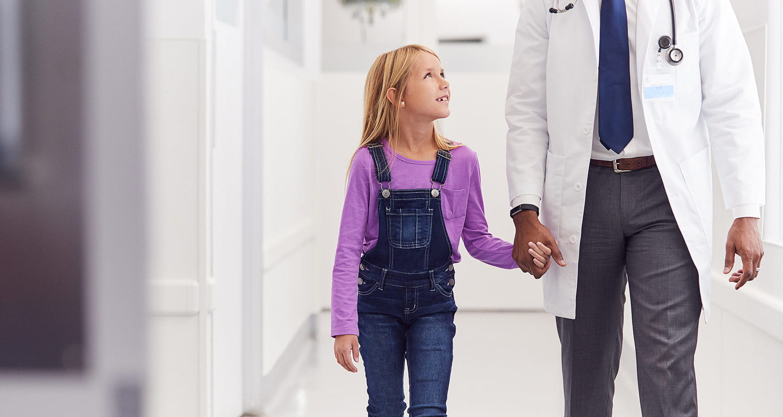 Close up of male paediatric doctor walking along hospital corridor holding hands with girl patient