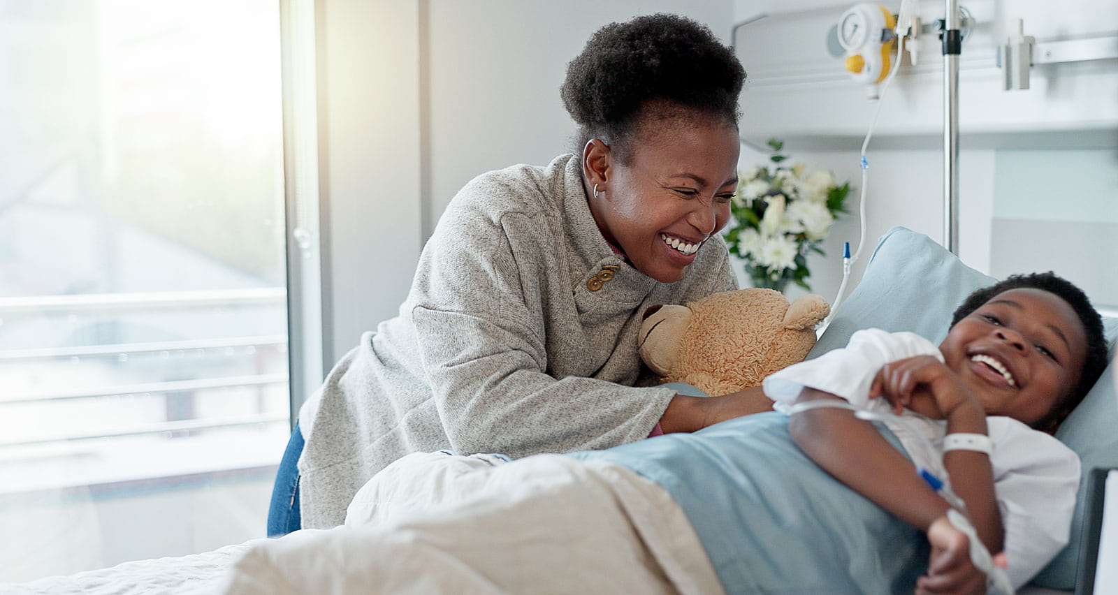 A young boy smiles in a hospital bed as his mother and doctor are looking in