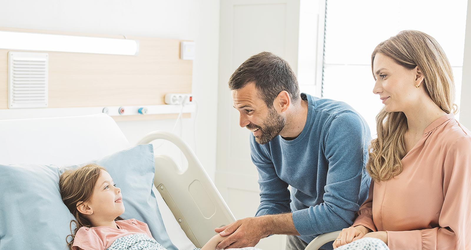 A little girl in a hospital bed while her mother and father are visiting