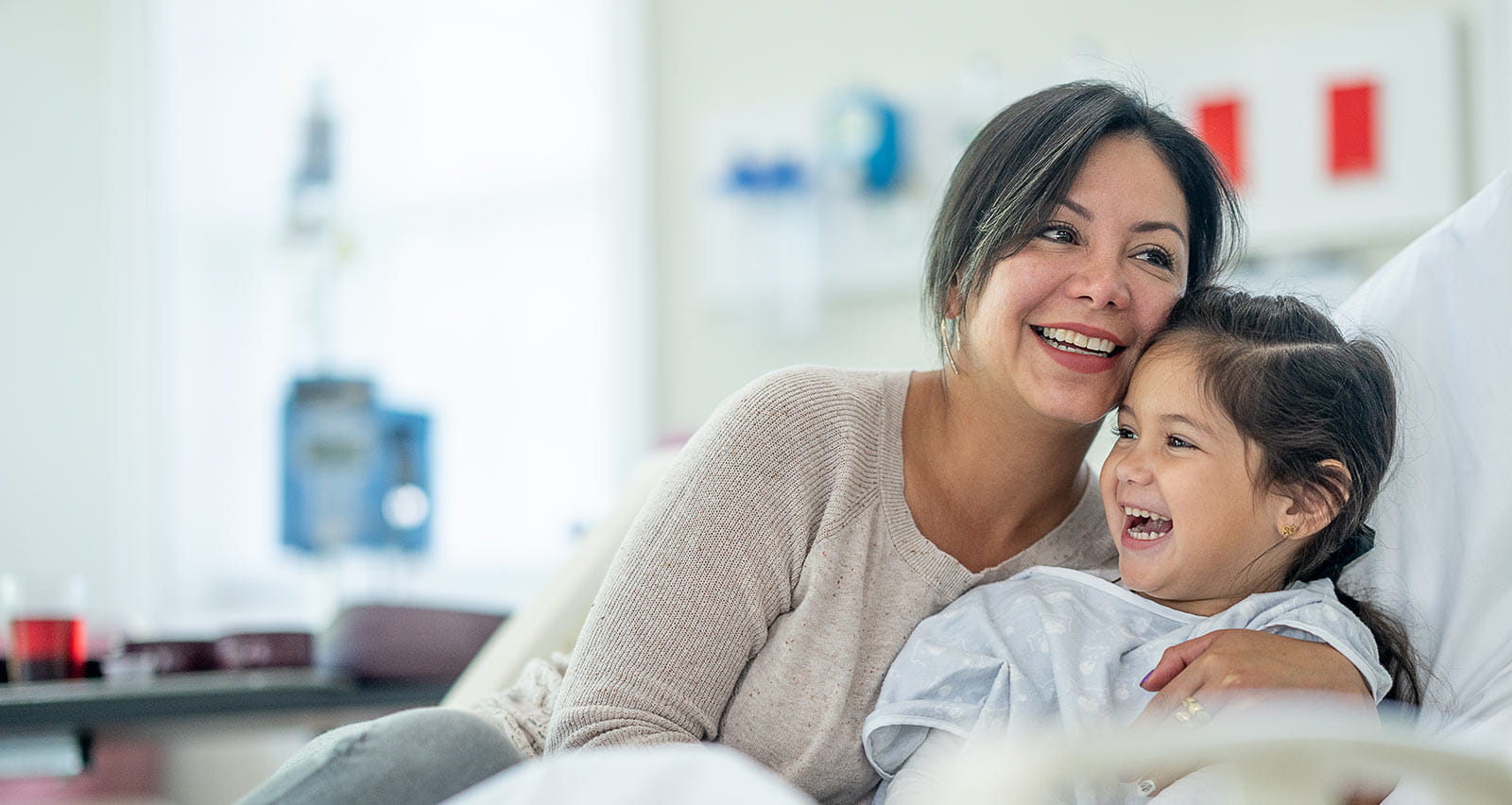 A mother sits up in a hospital bed with her daughter as she comforts and reassures her before surgery